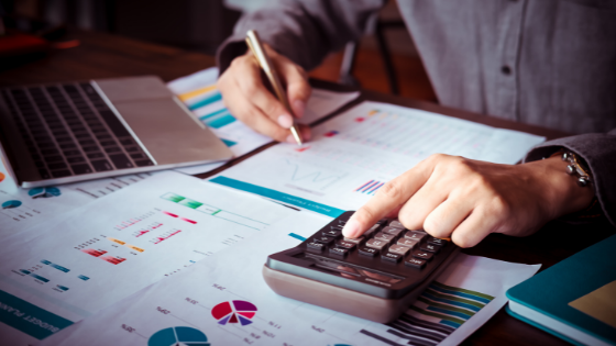 Someone writing on data and financial documents while tapping on a calculator; image used for blog post about selling trade or business property