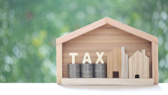 A photo of a wooden house frame filled with coins, smaller houses and the letters, "tax;" image used for blog post about saving income tax on estate plan