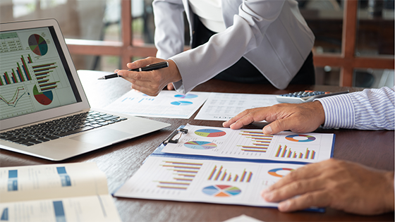 An image of two people looking at analytics on a laptop screen and data and graphics on loose-leaf paper; image used for blog post about year-end tax planning topics to discuss with a tax advisor to maximize savings