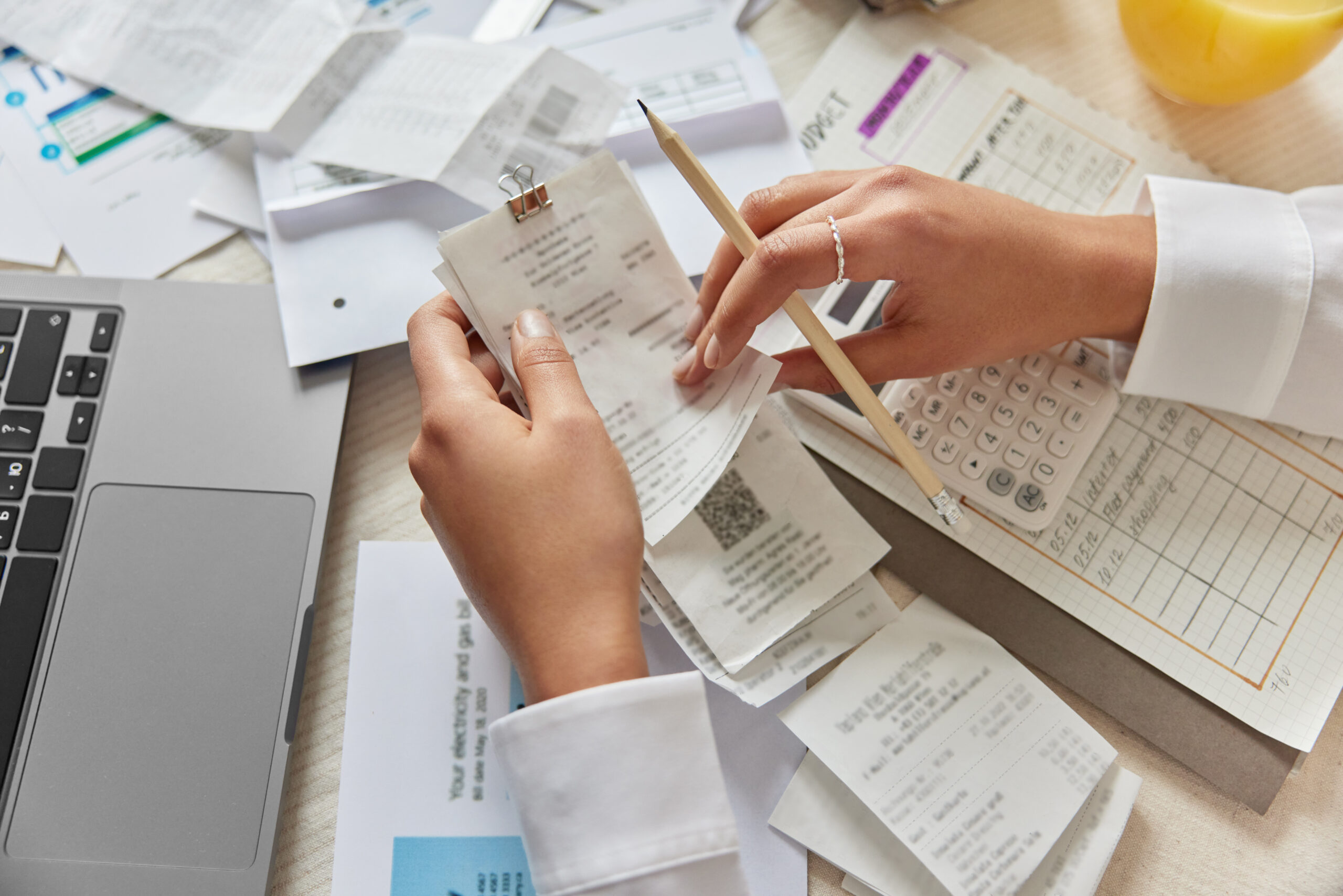 Unrecognizable woman holds receipts pencil uses laptop plans budget manages expenses calculates financial reports checks internet banking service poses at desktop with papers around. View from above